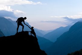 Silhouette of two people helping each other on top of a mountain.